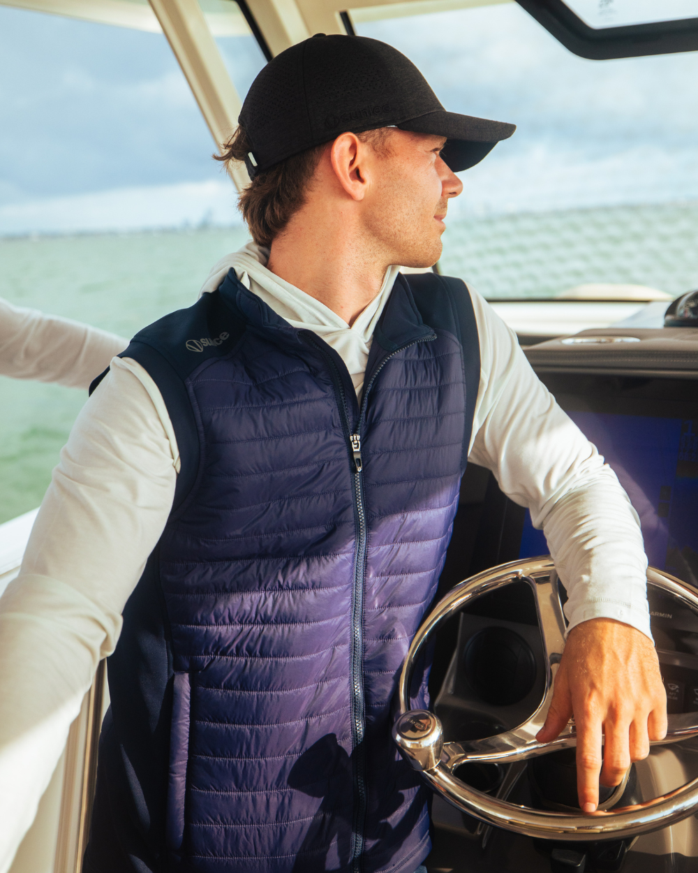 Man wearing a navy Sunice water-resistant performance vest and long-sleeve top while steering a boat on open water.