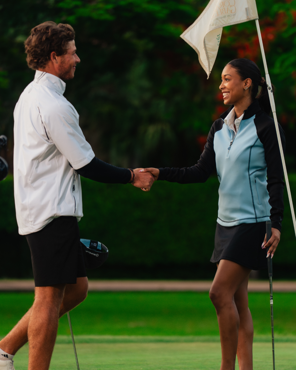 Two golfers smiling and shaking hands near the flagstick on a lush green, wearing Sunice lightweight golf outerwear.