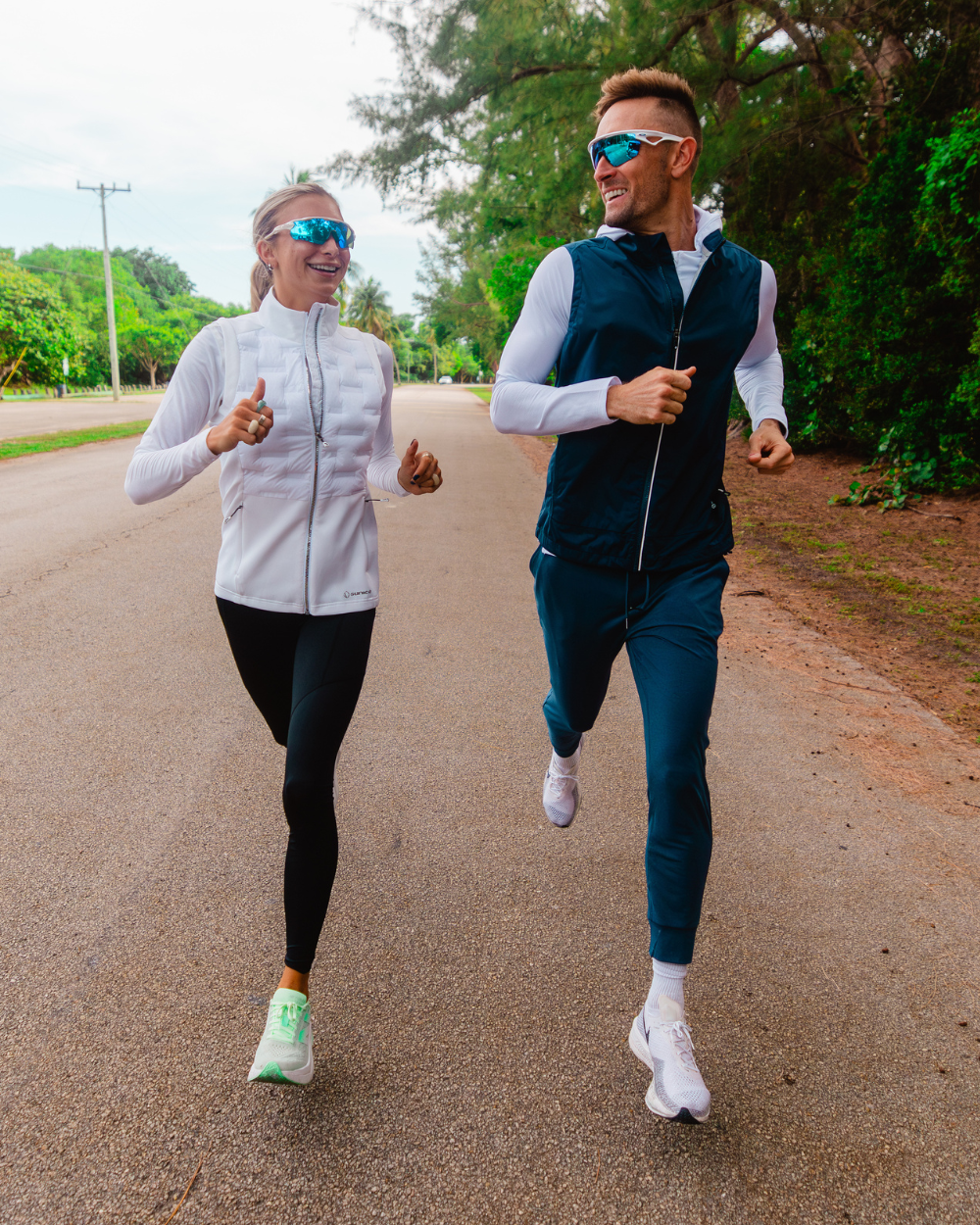 Two runners smiling while jogging on a tree-lined road, wearing Sunice performance vests and athletic apparel.