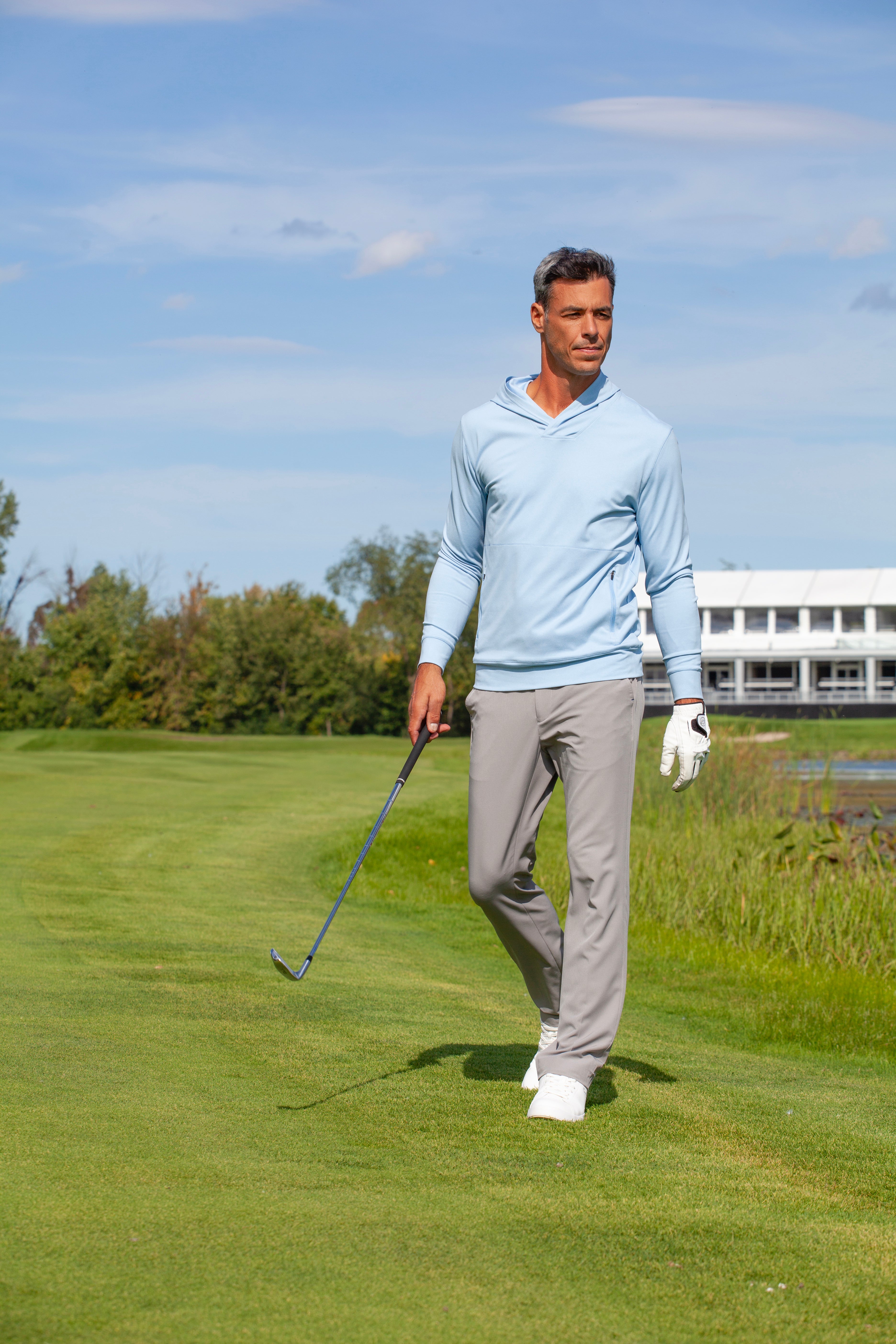 Man on golf course wearing Sunice Adam Aerosoft stretch pullover hoodie in blue bell melange and grey trousers.
