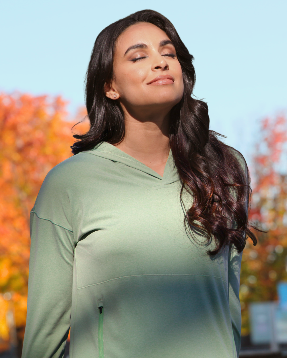 Woman wearing a mint green Sunice performance pullover hoodie outdoors with vibrant fall foliage in the background.