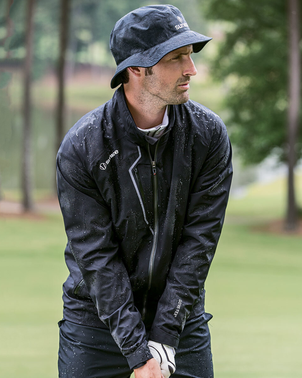 Man wearing a black Sunice waterproof golf jacket and bucket hat playing in light rain on a golf course.