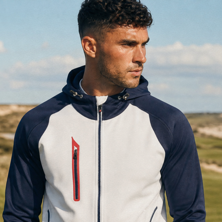 Model wearing a white and navy Sunice hooded jacket with red zipper detail at a coastal golf course.