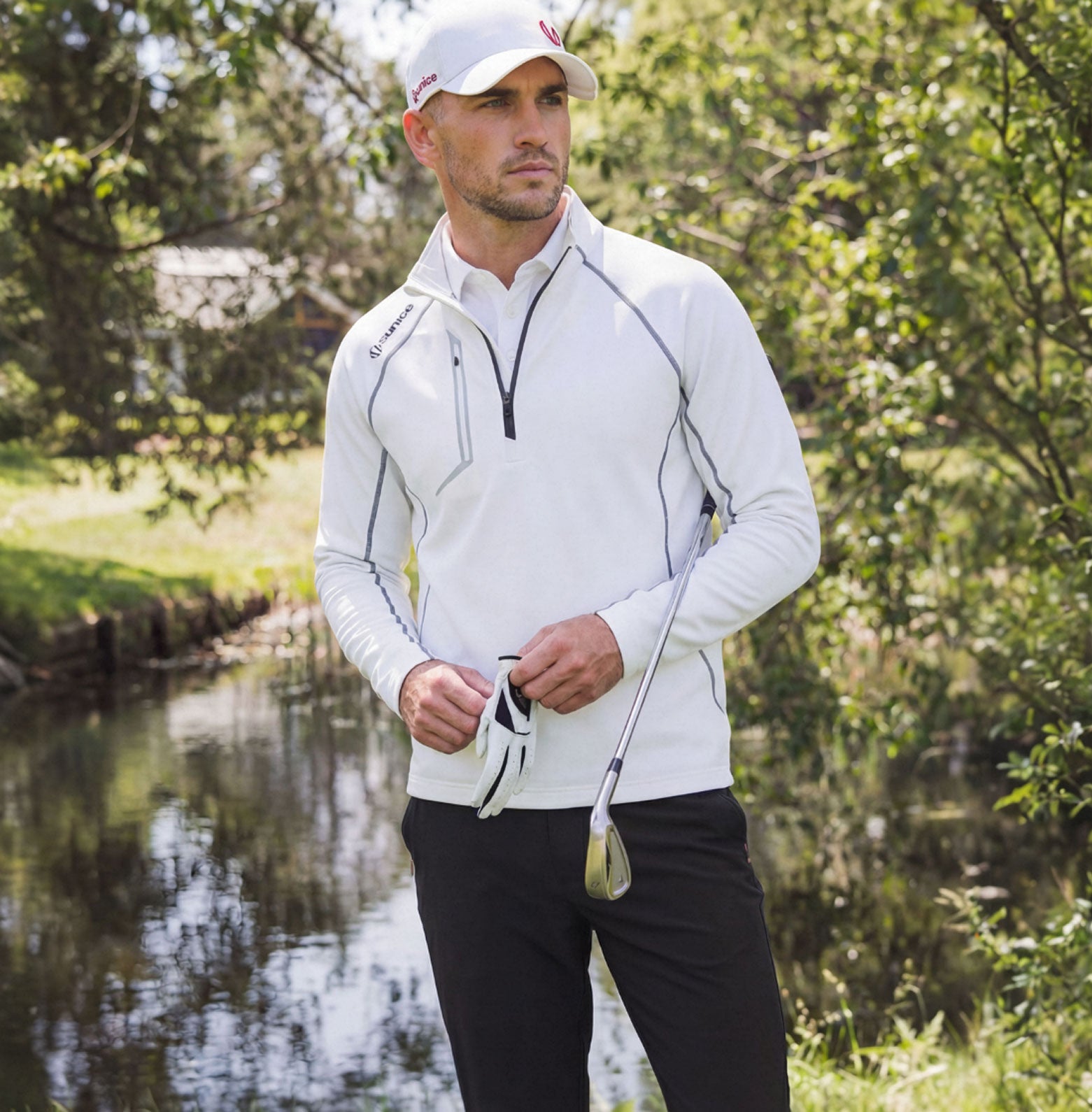 Man in a white jacket and black pants holding a golf club by a body of water with trees in the background