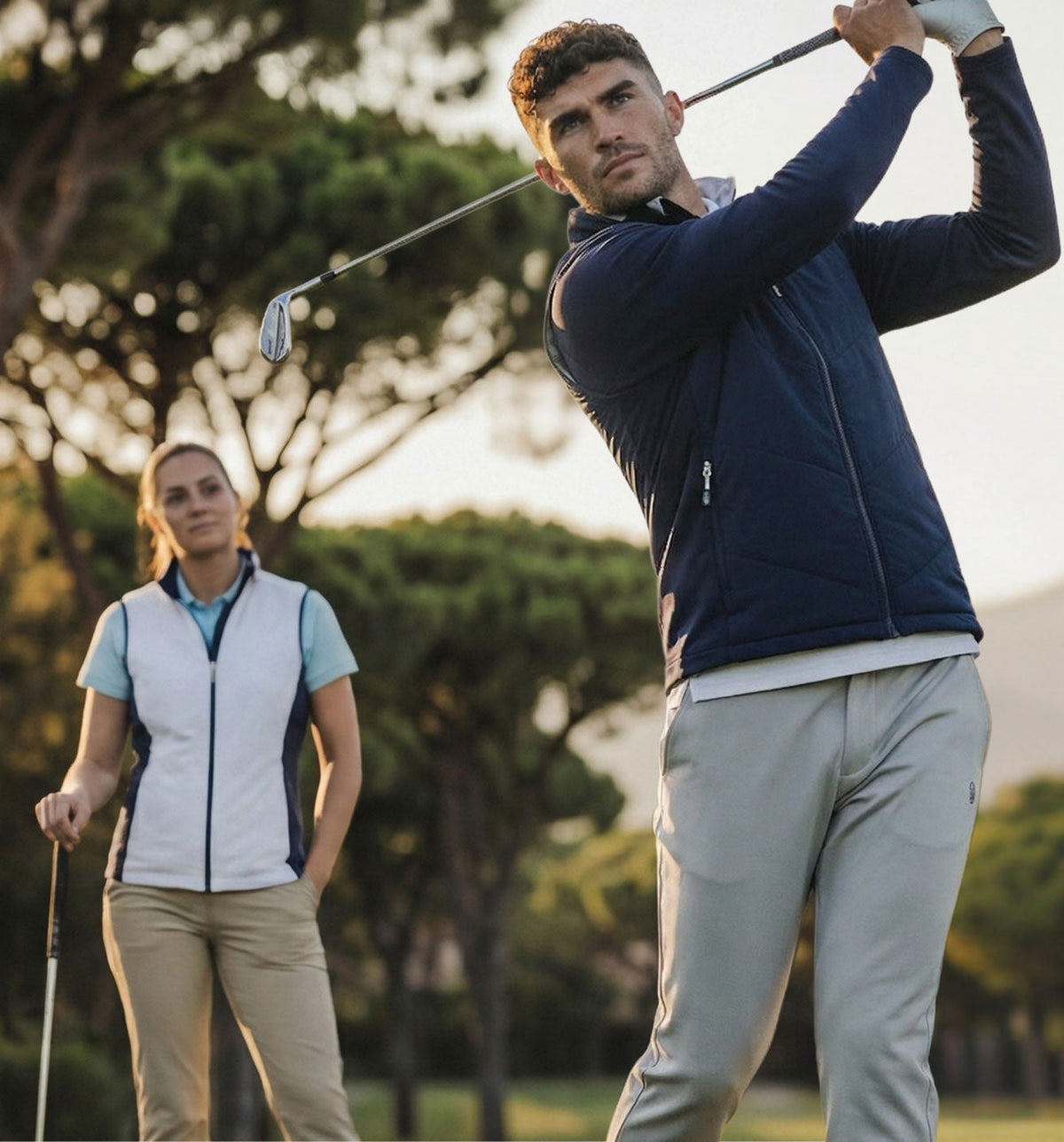 Man swinging a golf club with a woman standing behind him on a golf course.
