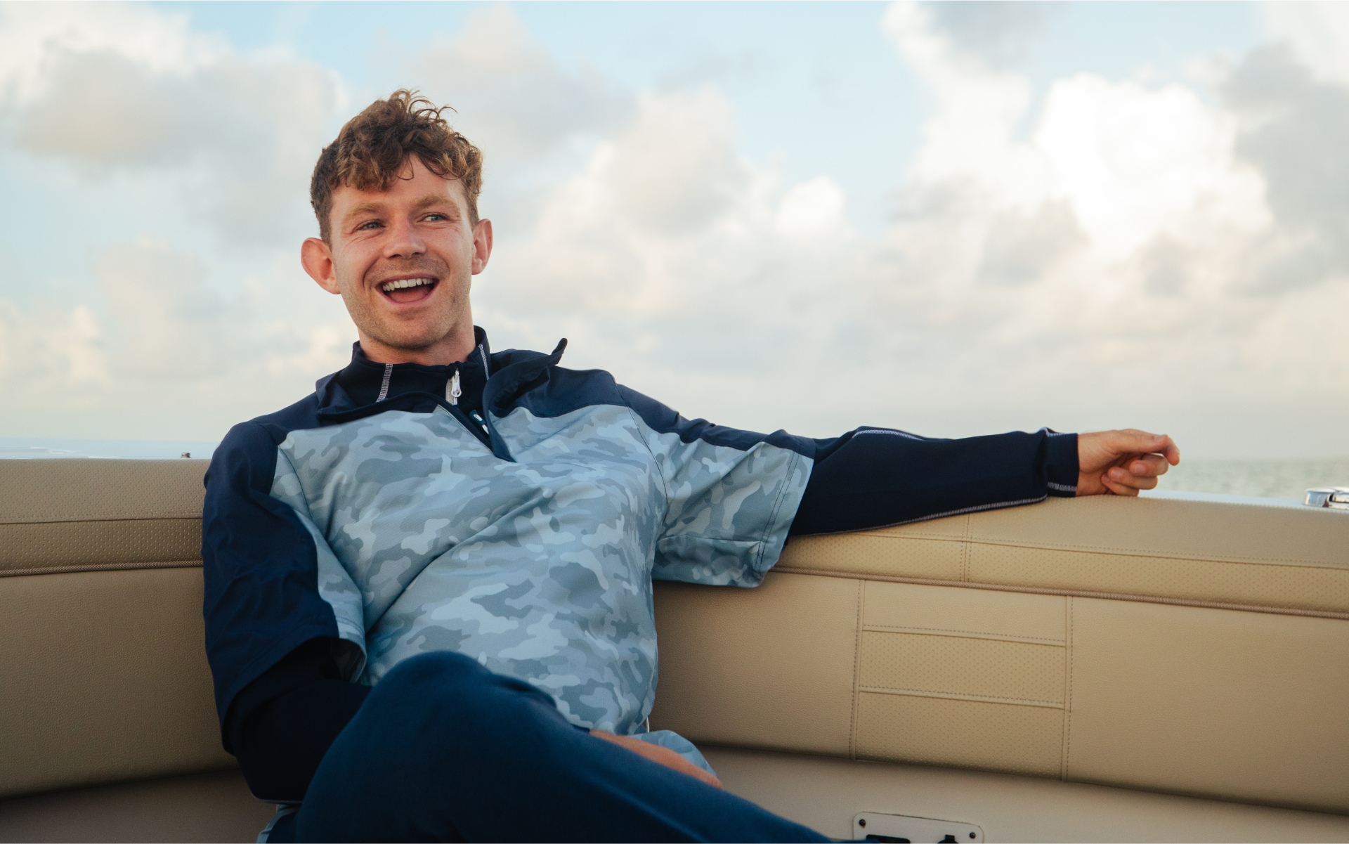 Man sitting on a boat deck with a two tone blue patterned Sunice quarter-zip, smiling.